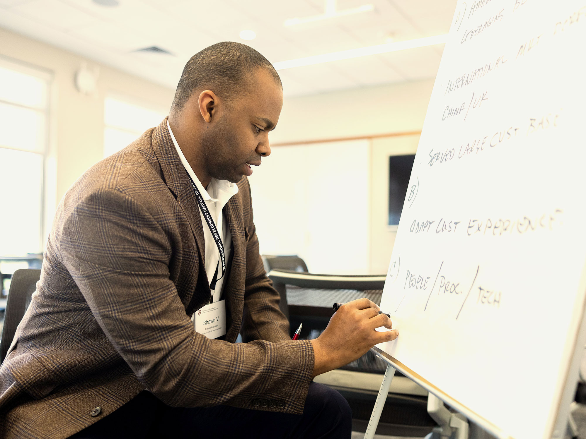 Man in a brown blazer writing key points on a whiteboard during a strategy session.