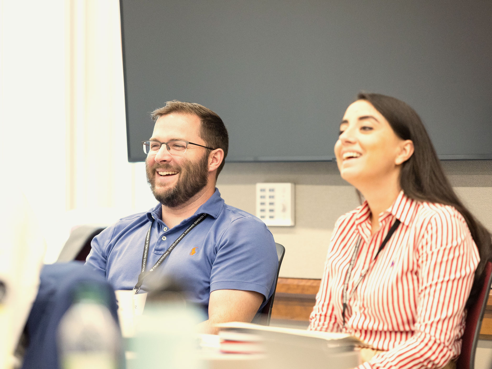 Two adult learners in a classroom smiling and laughing while listening to a speaker out of frame.