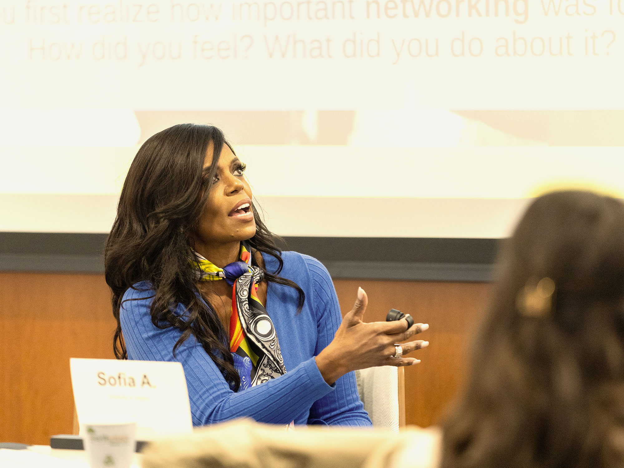 Woman in a blue top and colorful scarf making a point during a panel discussion.