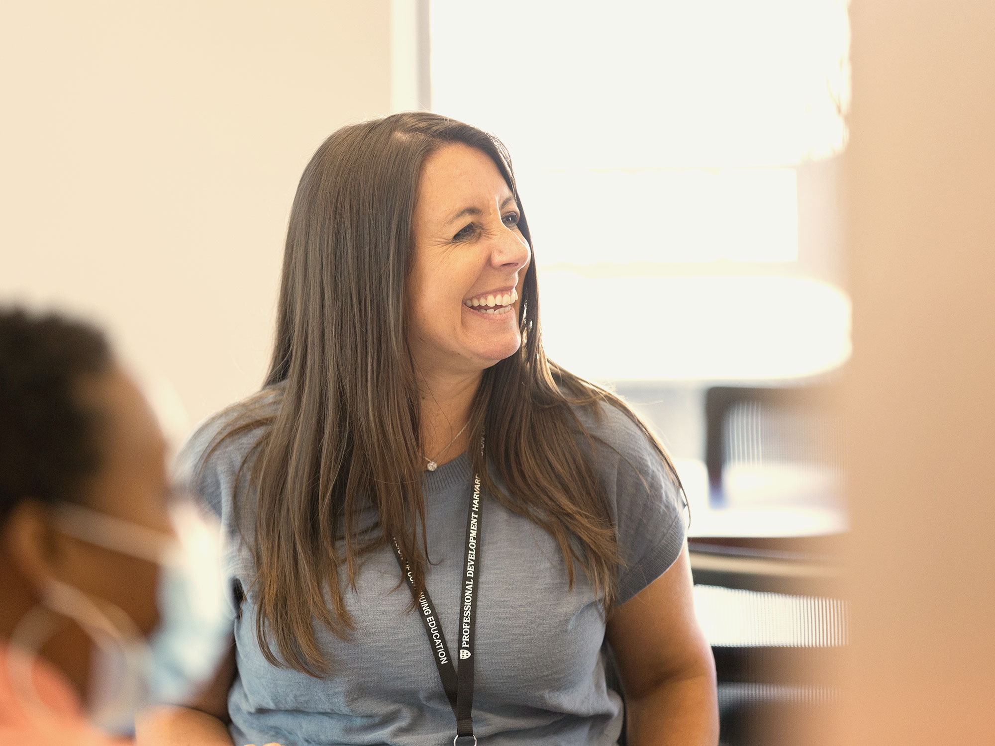 Woman in a gray shirt smiling and engaged during a classroom discussion.