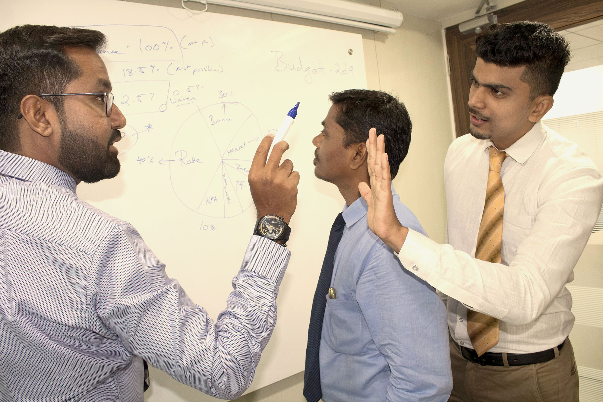 One man looks at a whiteboard while two men argue behind him.