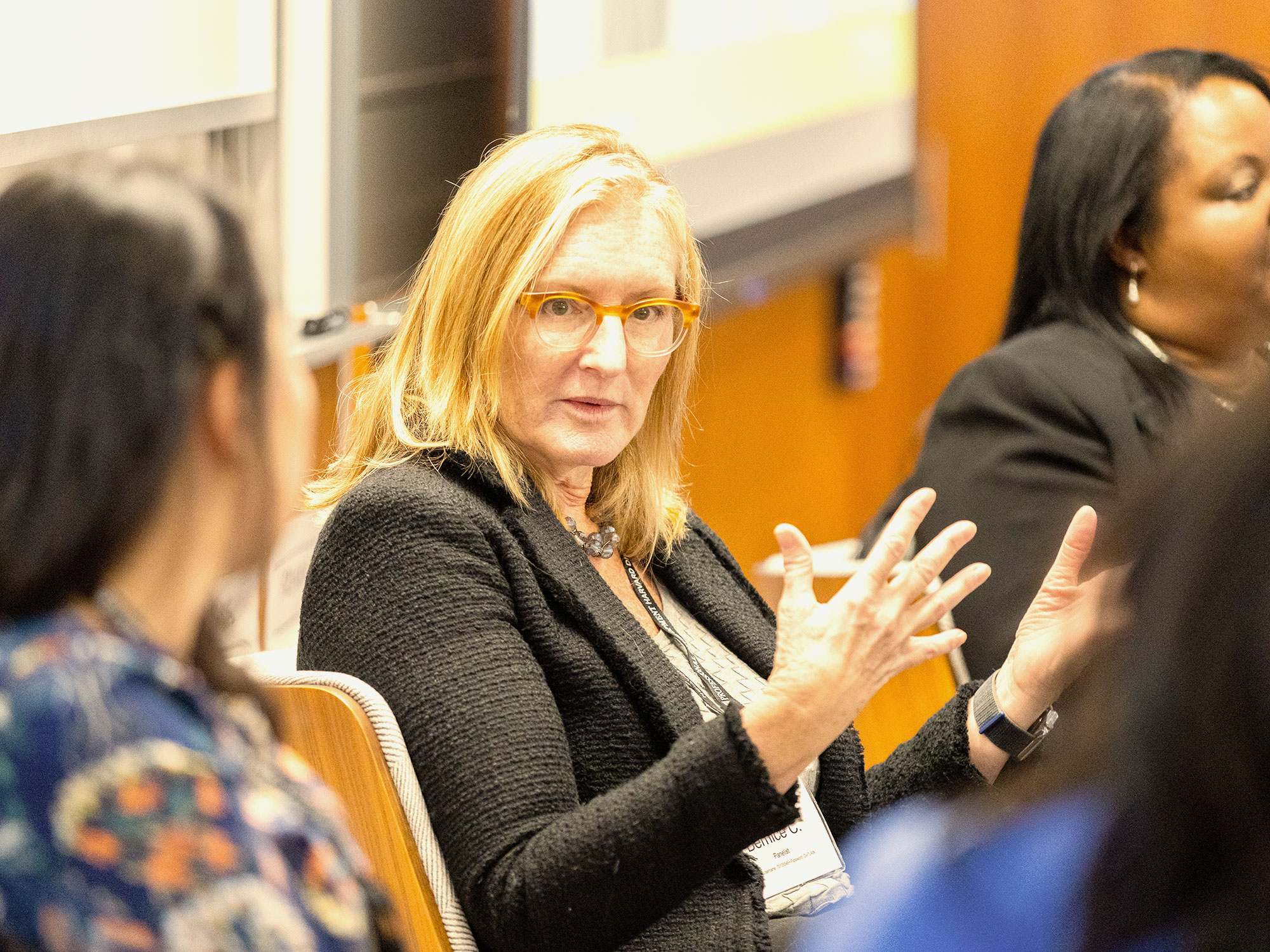 A woman in a black blazer speaking with her hands to a panel of women in professional attire.